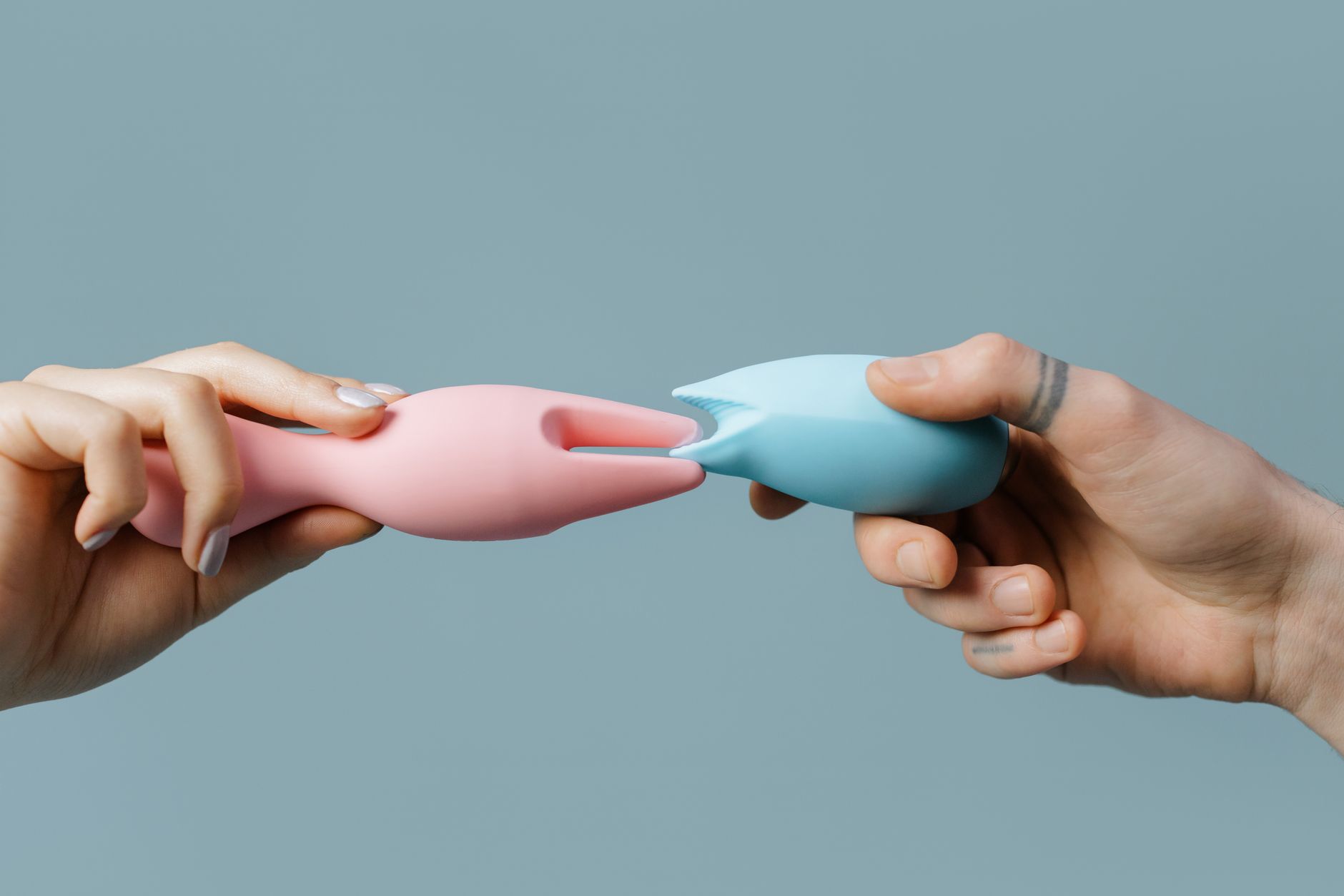Two hands holding pink and blue silicone clitoral vibrators against a pastel background