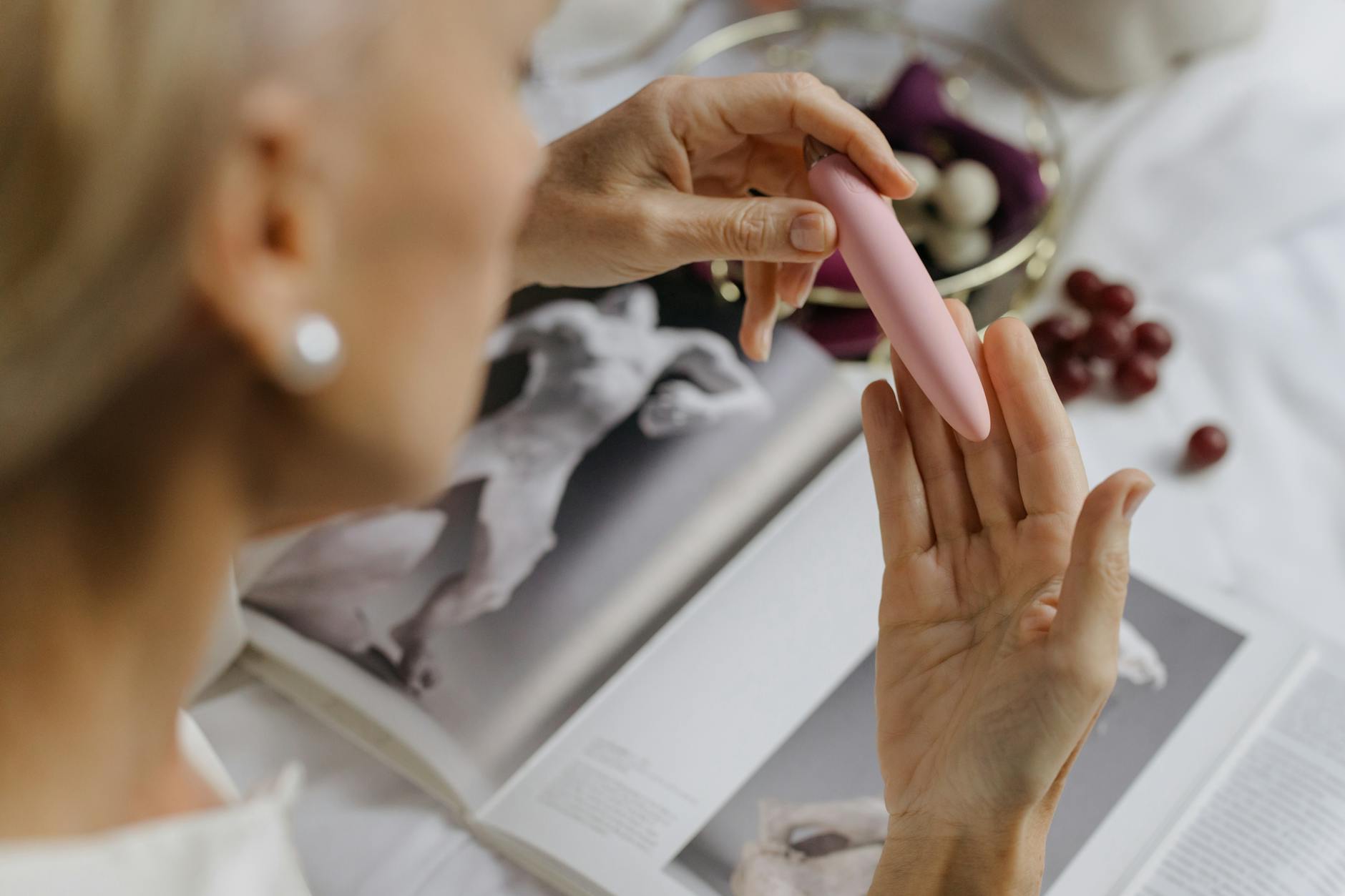 Close-up of a woman holding a pink vibrator, with art books nearby, emphasizing intimate personal pleasure.