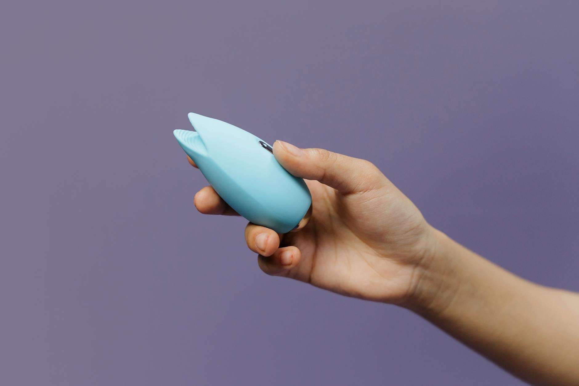 A blue silicone clitoral vibrator held in hand against a purple background, representing gentle pleasure and self-exploration