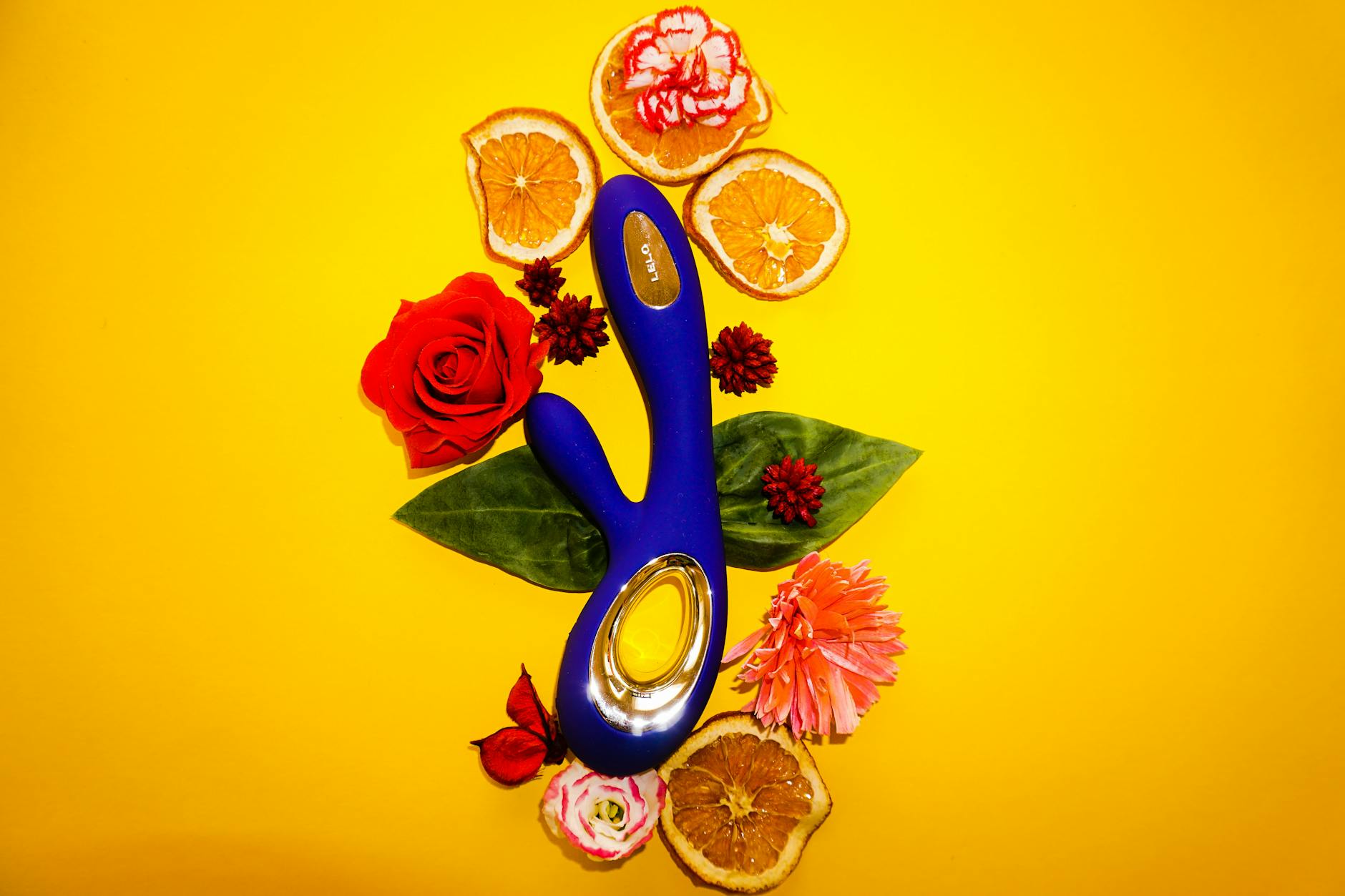 A blue lemon vibrator surrounded by fresh citrus and flowers on a bright yellow surface.